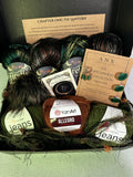 A curated yarn gift box from ANX Yarns featuring multiple skeins of yarn in forest green, black, and brown, along with a brown yarn label reading 'YarnArt ALLEGRO', a card titled 'The Witchwood Collection Chapter One', and a small card titled 'Chapter One: The Lantern'. The items are arranged in a black box lined with green yarn, set against a light green background.
