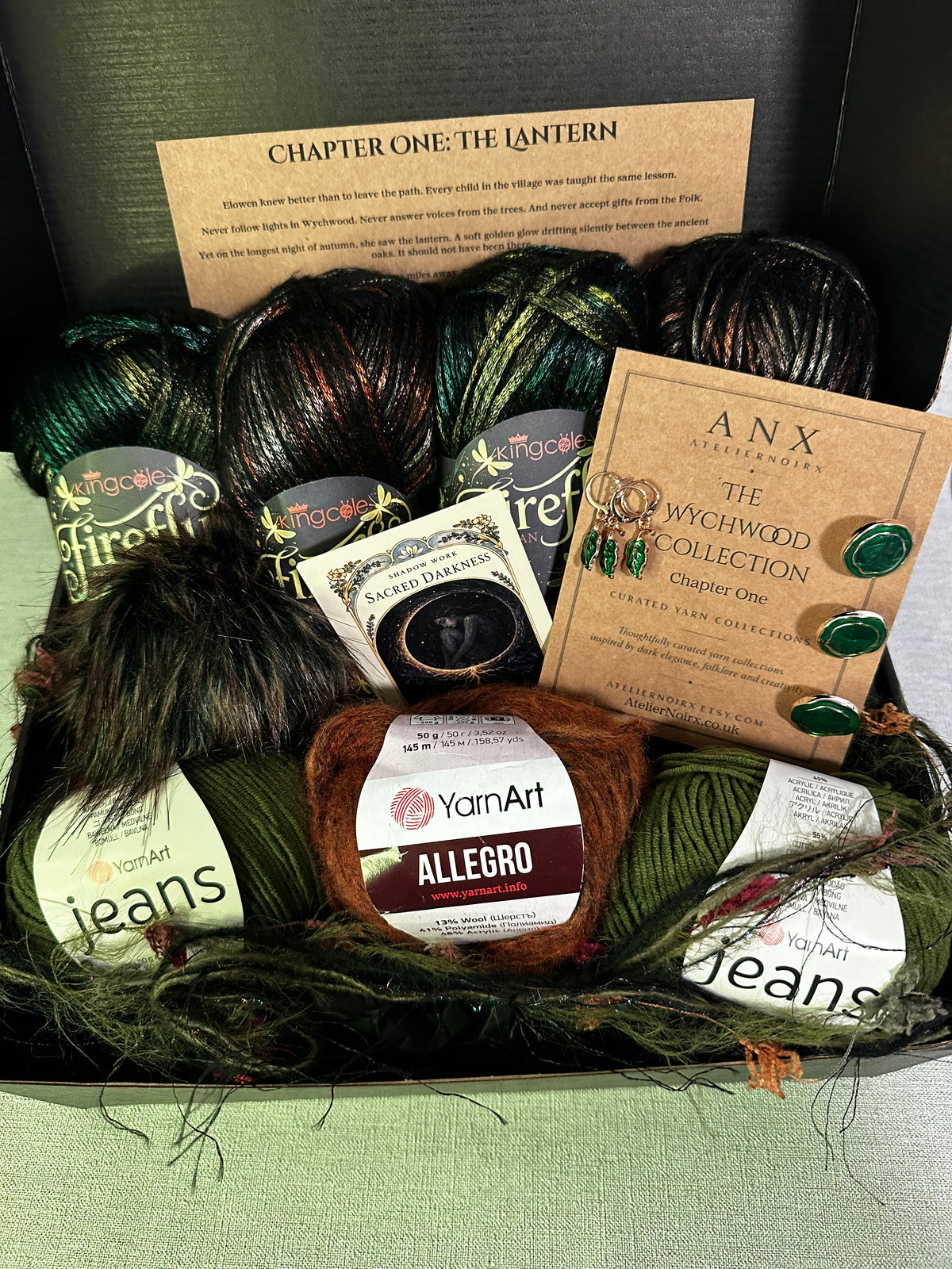 A curated yarn gift box from ANX Yarns featuring multiple skeins of yarn in forest green, black, and brown, along with a brown yarn label reading 'YarnArt ALLEGRO', a card titled 'The Witchwood Collection Chapter One', and a small card titled 'Chapter One: The Lantern'. The items are arranged in a black box lined with green yarn, set against a light green background.